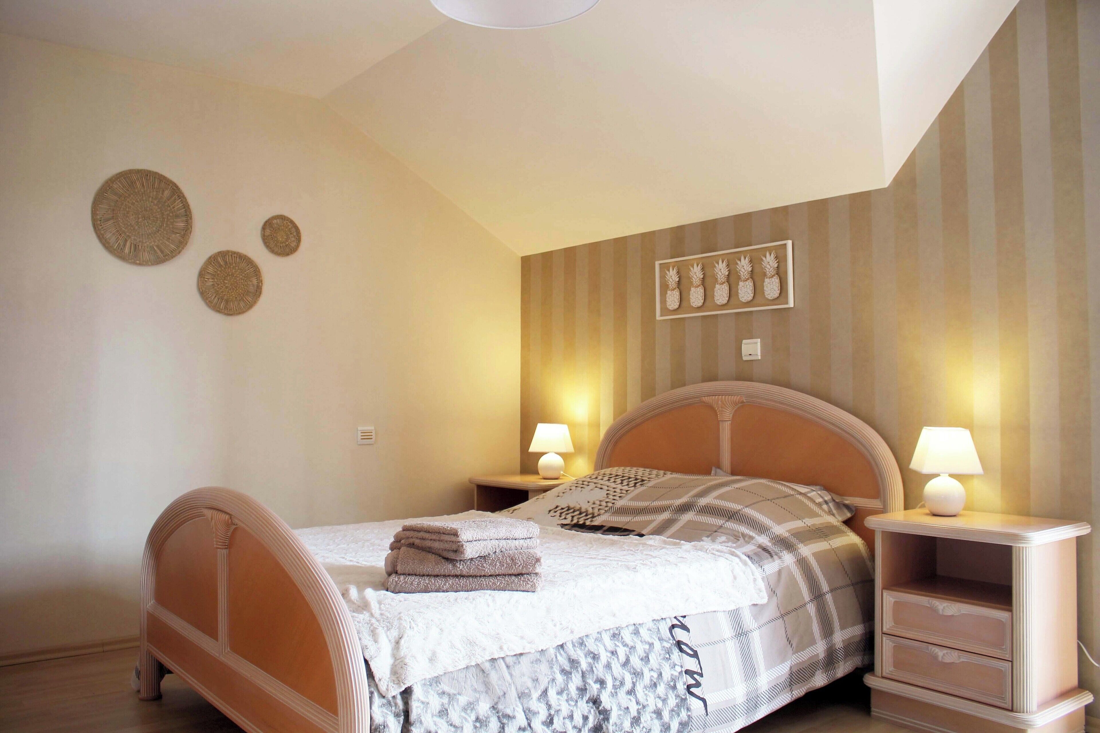 Una cama bien tendida con una colcha blanca y estampada, dos lámparas de noche y una mesita de noche con lámpara.