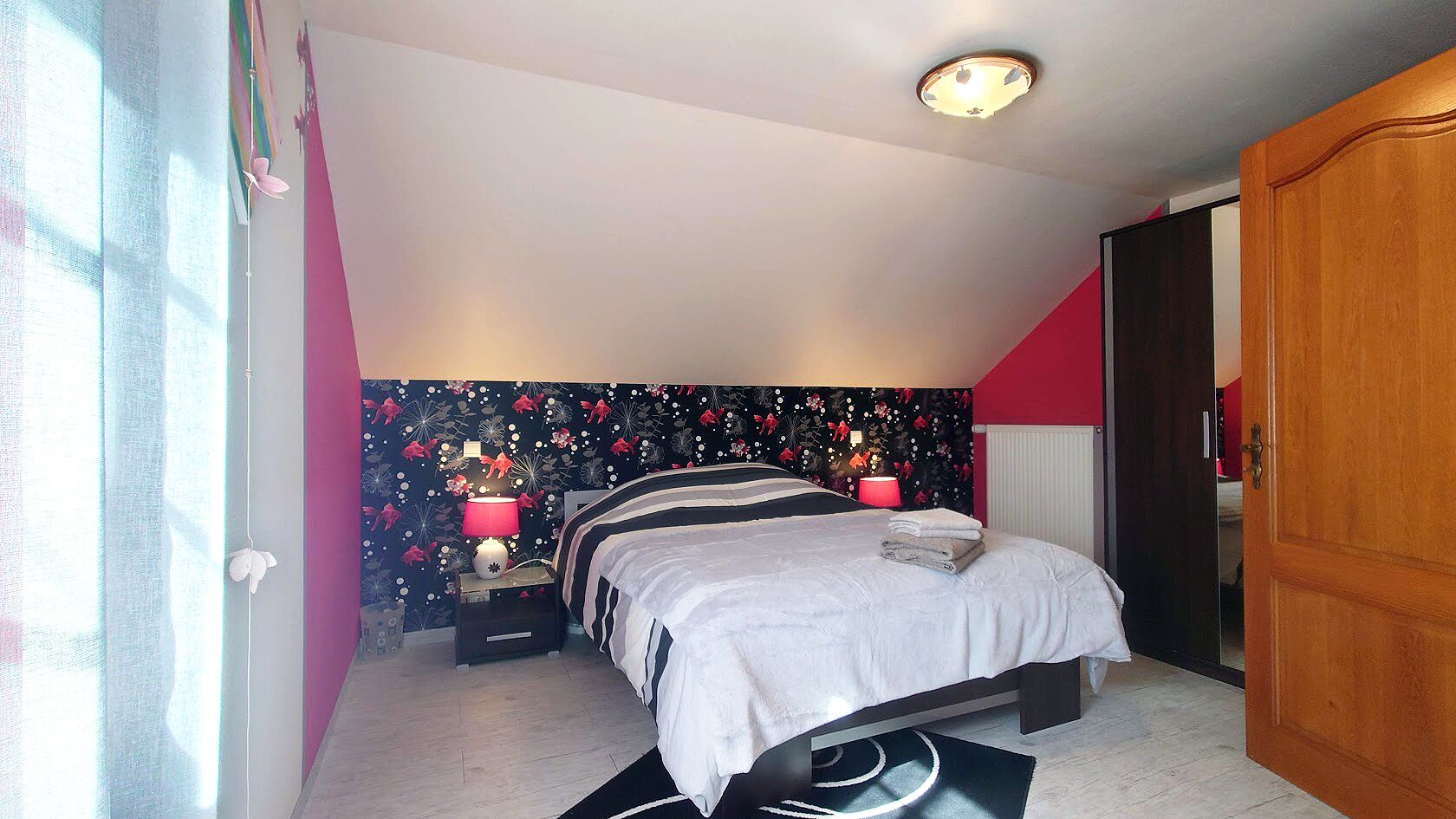 Une chambre à coucher comprenant un lit, des tables de chevet, une commode, un mur recouvert d’un papier peint à motifs floraux, un luminaire au plafond et une porte.