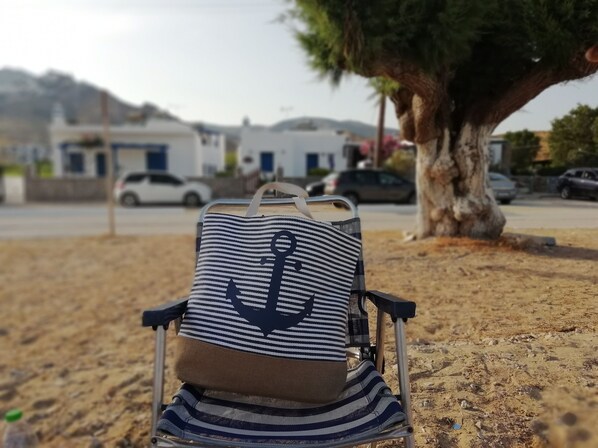 Una silla de playa con tela a rayas y un diseño de ancla, ubicada sobre arena, con un árbol y coches aparcados al fondo.