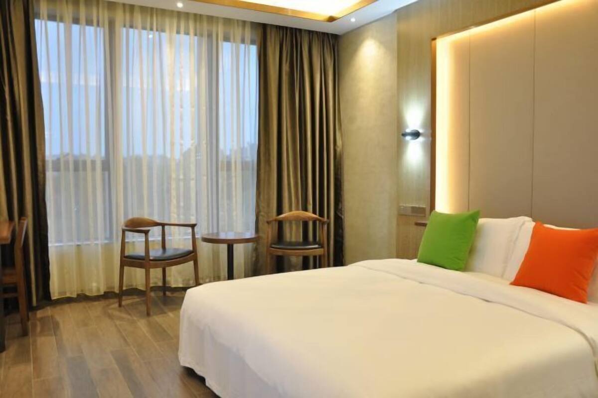 Habitación de hotel con una cama grande, dos sillas, una mesita y un ventanal con cortinas.