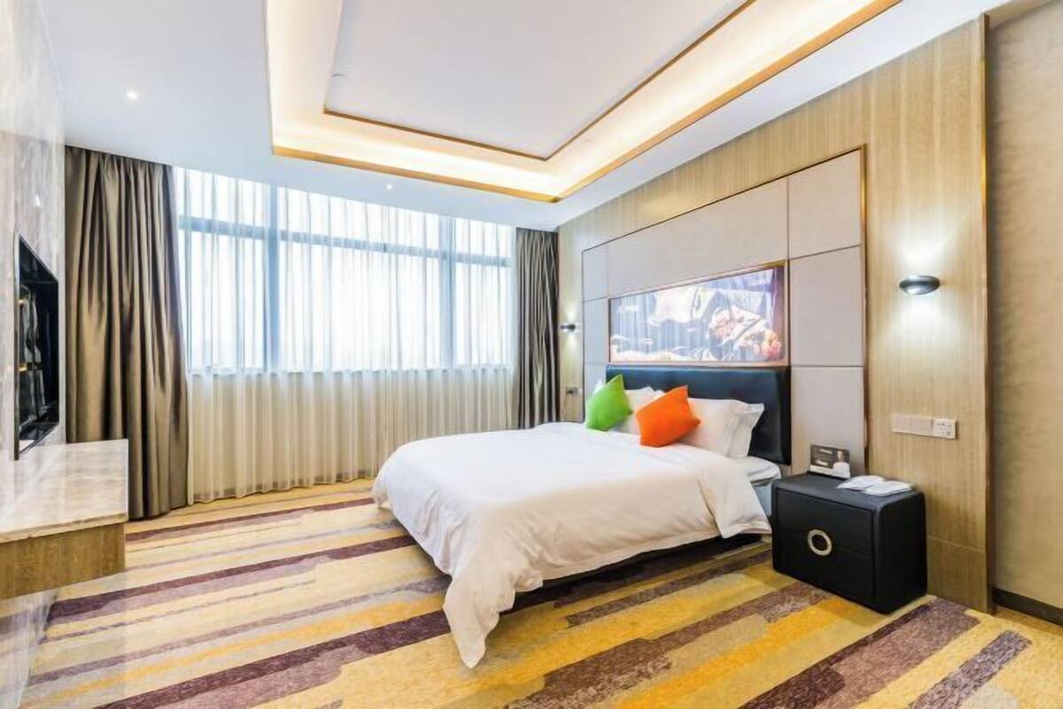 Una habitación de hotel moderna con una cama grande, un televisor de pantalla plana, una mesita de noche y un ventanal con cortinas.