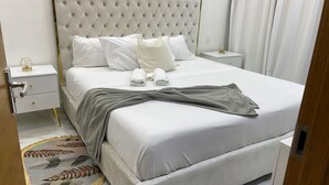 Ein ordentlich gemachtes Bett mit weißen Leinentüchern und einer grauen Tagesdecke, ein gepolstertes Kopfende sowie ein Nachttisch mit Lampe und einem Glasobjekt.