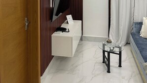 Ein modernes Hotelzimmer mit Holztür, einem flachbildfernseher an einer weißen Konsole, einem kleinen runden Tisch mit Glasplatte und einem blauen Sofa mit weißen Kissen.