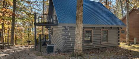 A log cabin with a balcony, a gravel path, and a forested background.