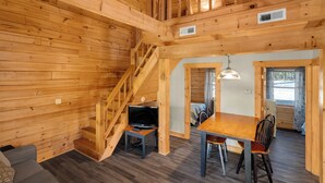 Interior de una cabaña de madera con comedor, una escalera y un televisor sobre una mesita.