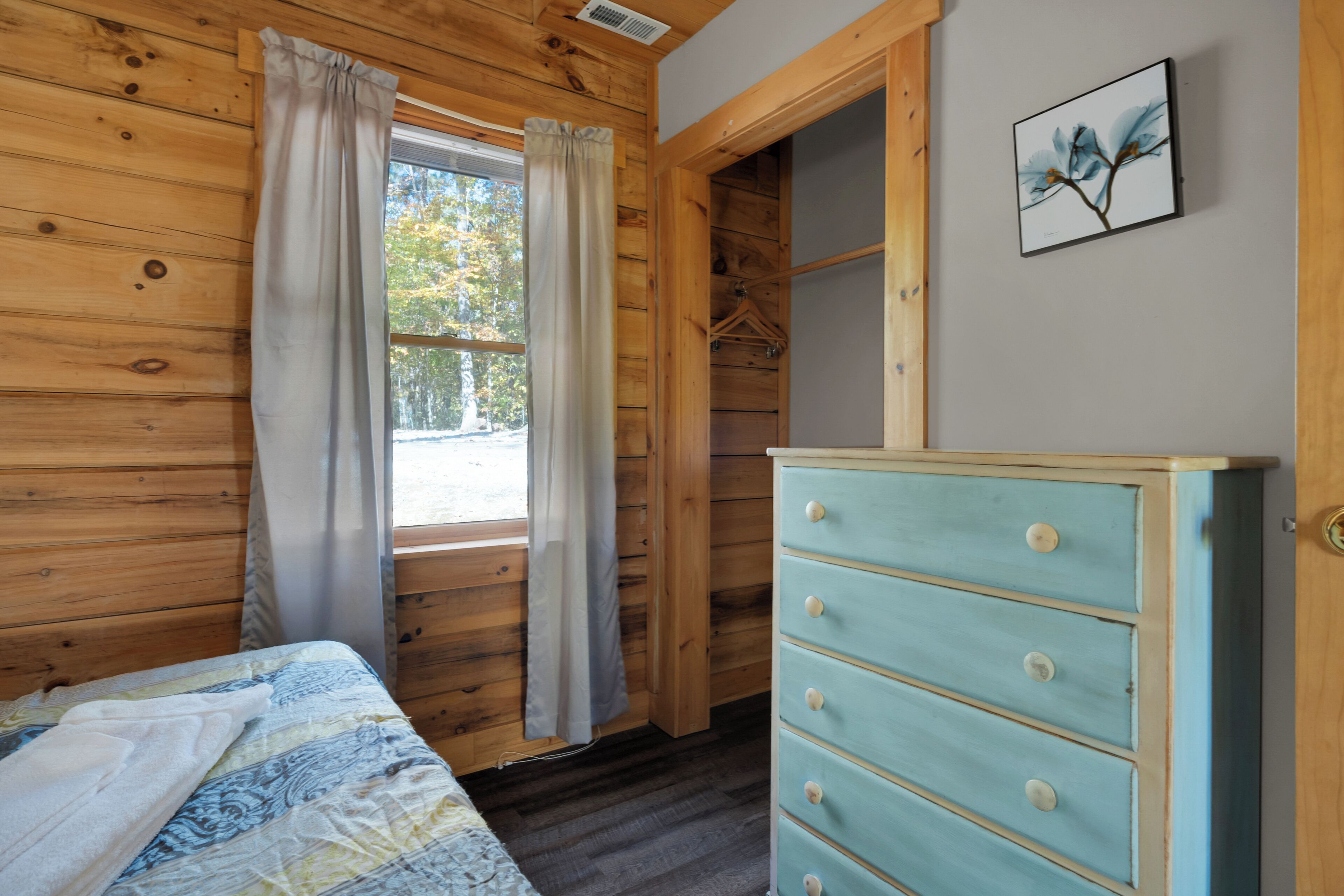 A wooden cabin room with a bed, a dresser, and a window with curtains.