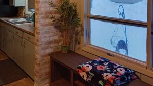 Ein gemütliches Zimmer mit Holzbalkendecke, einer Wand in Ziegeloptik, einem Fenster mit Blick auf ein Haus, einer Bank mit Blumenkissen und einer kleinen Topfpflanze.