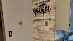 Ein kleiner Essbereich mit einem runden Holztisch und zwei weißen Plastikstühlen. Dahinter eine Backsteinwand mit dem Schriftzug 'gather'.
