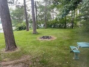 Relax in a serene grassy area surrounded by tall trees, featuring a fire pit, hammock, and dining space.