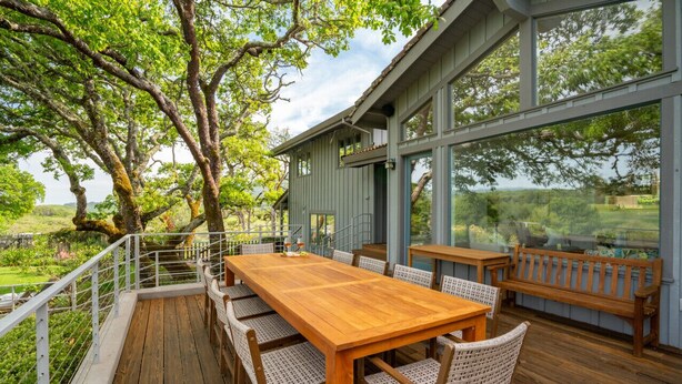 A wooden deck with a dining table and chairs, surrounded by large windows offering a view of trees and greenery.