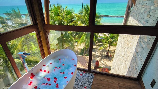 Una bañera grande llena de agua y pétalos de rosa, ubicada cerca de una ventana con vistas a una playa y palmeras.