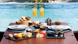 Una mesa dispuesta con diversos productos para el desayuno, como pasteles, frutas y bebidas, con vista a una piscina, una playa y palmeras al fondo.