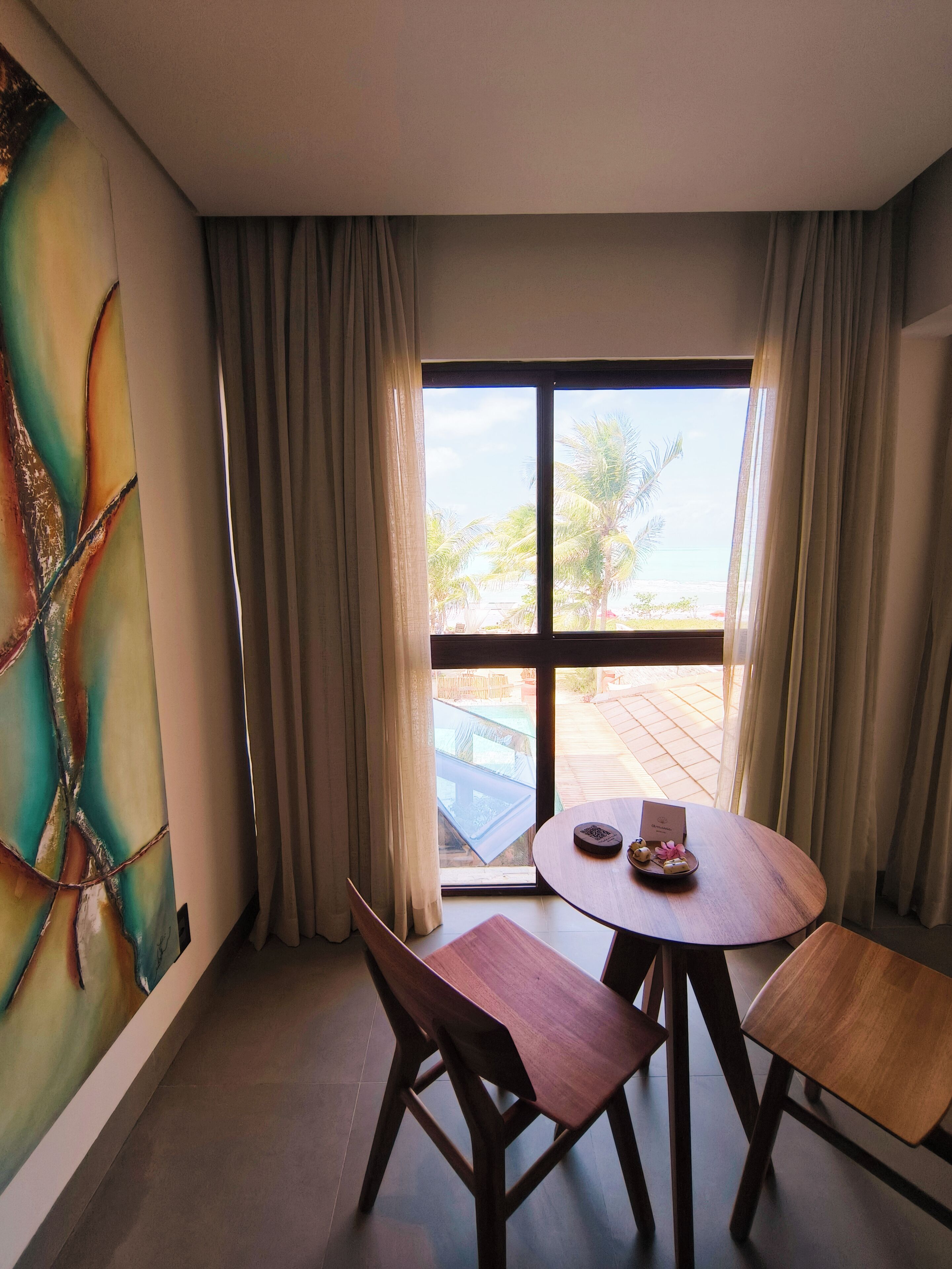 A room with a large window, a wooden table with chairs, and a colorful abstract painting on the wall.