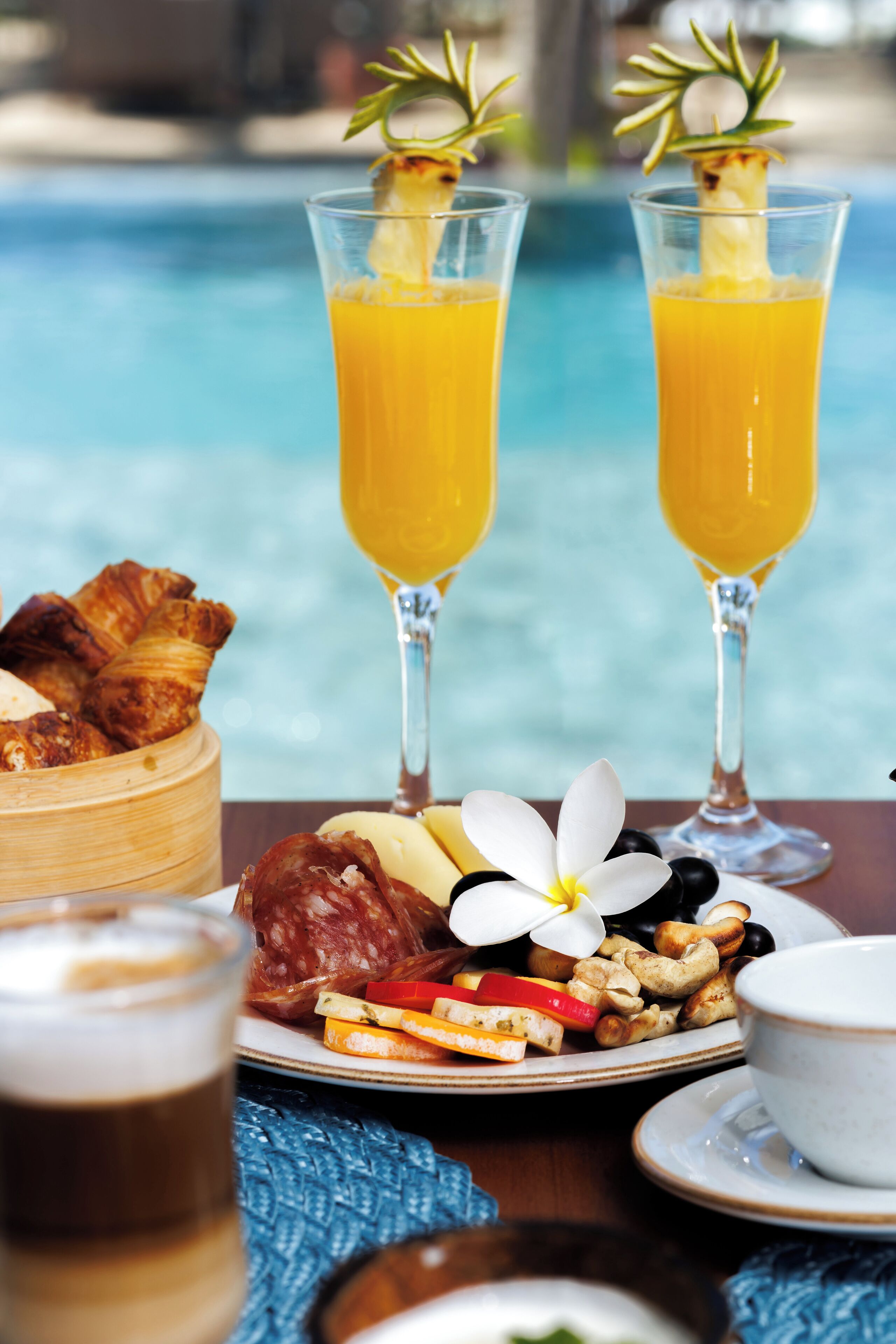 A table with breakfast items including croissants, a plate of fruits, nuts, and a cup of coffee, with a pool in the background.