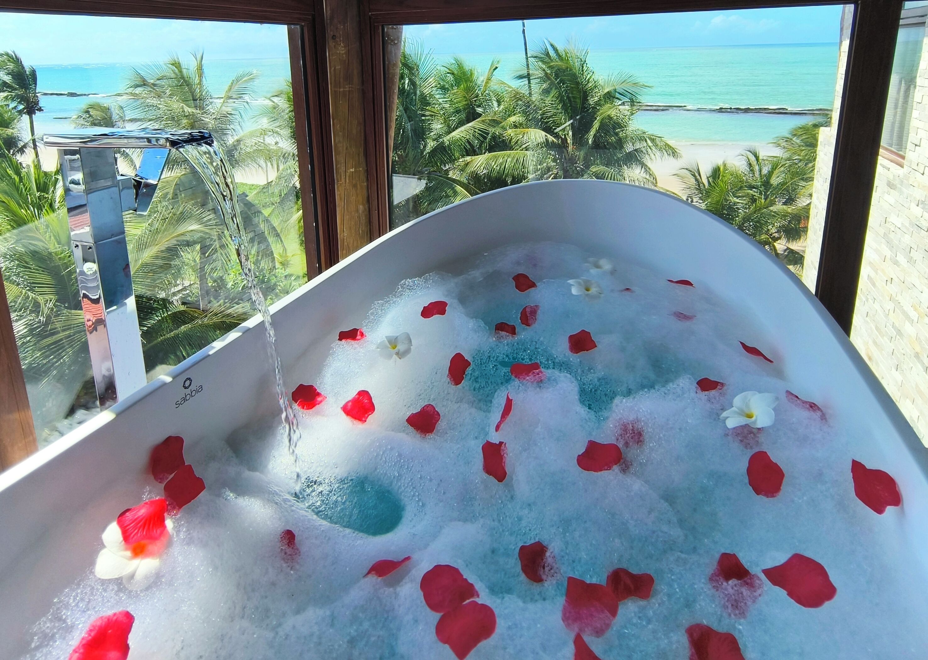 Eine Badewanne voller Blasen und Rosenblätter, mit Blick auf einen Strand und Palmen durch ein Fenster.