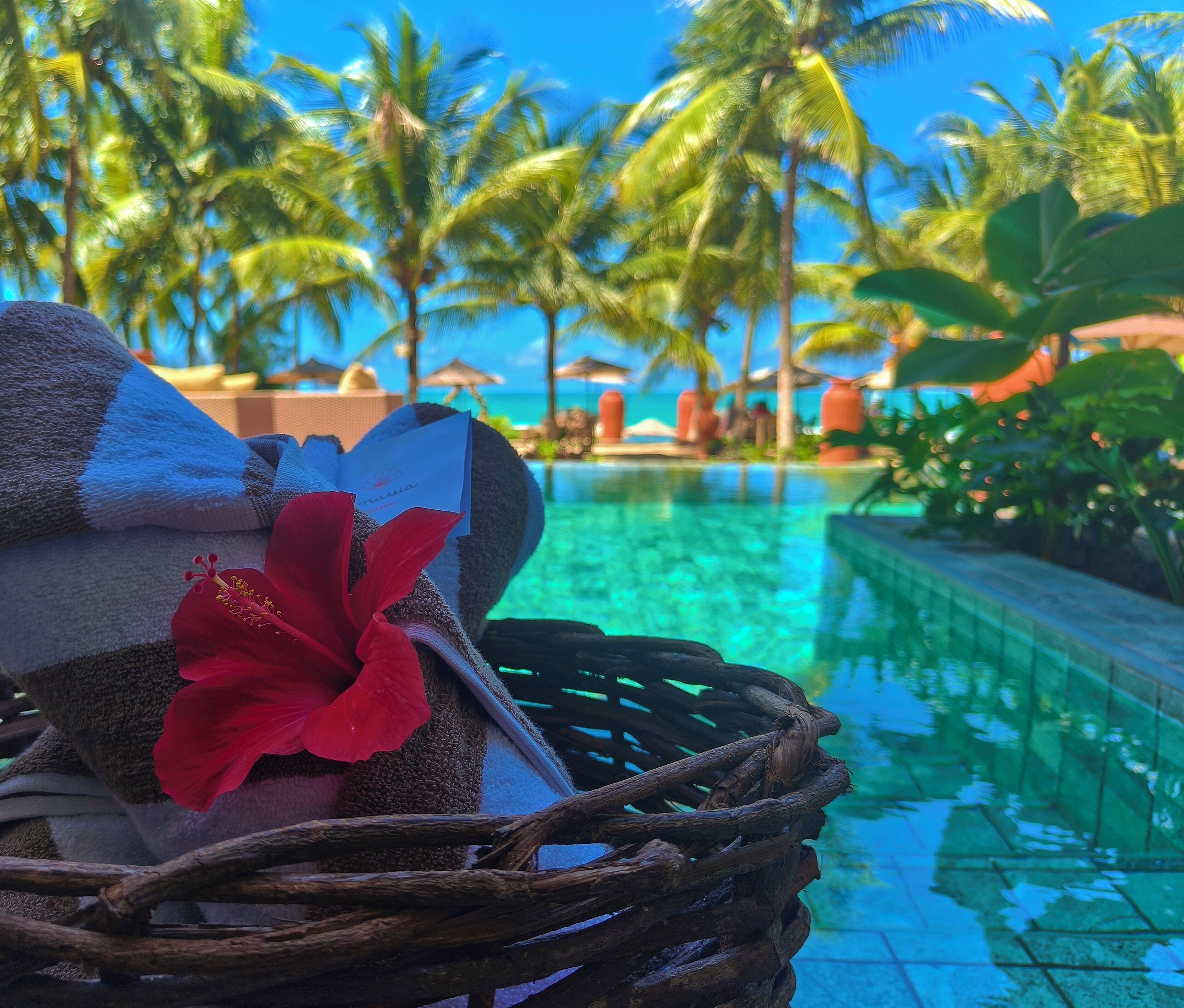 A wicker basket with towels and a red flower, overlooking a pool with palm trees and a clear blue sky.