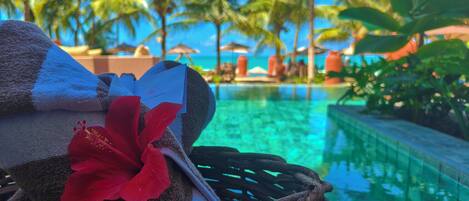 A wicker basket with towels and a red flower, overlooking a pool with palm trees and a clear blue sky.