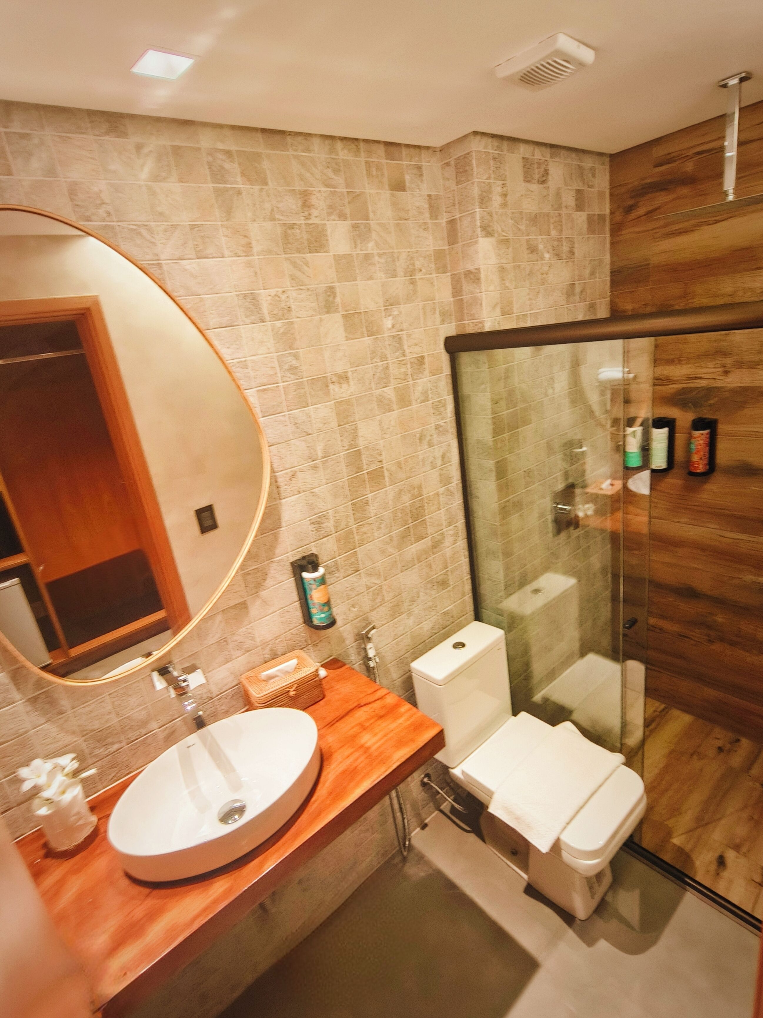 Een moderne badkamer met een houten wastafelmeubel, een ronde spiegel, een toilet en een douchecabine met glazen wanden.