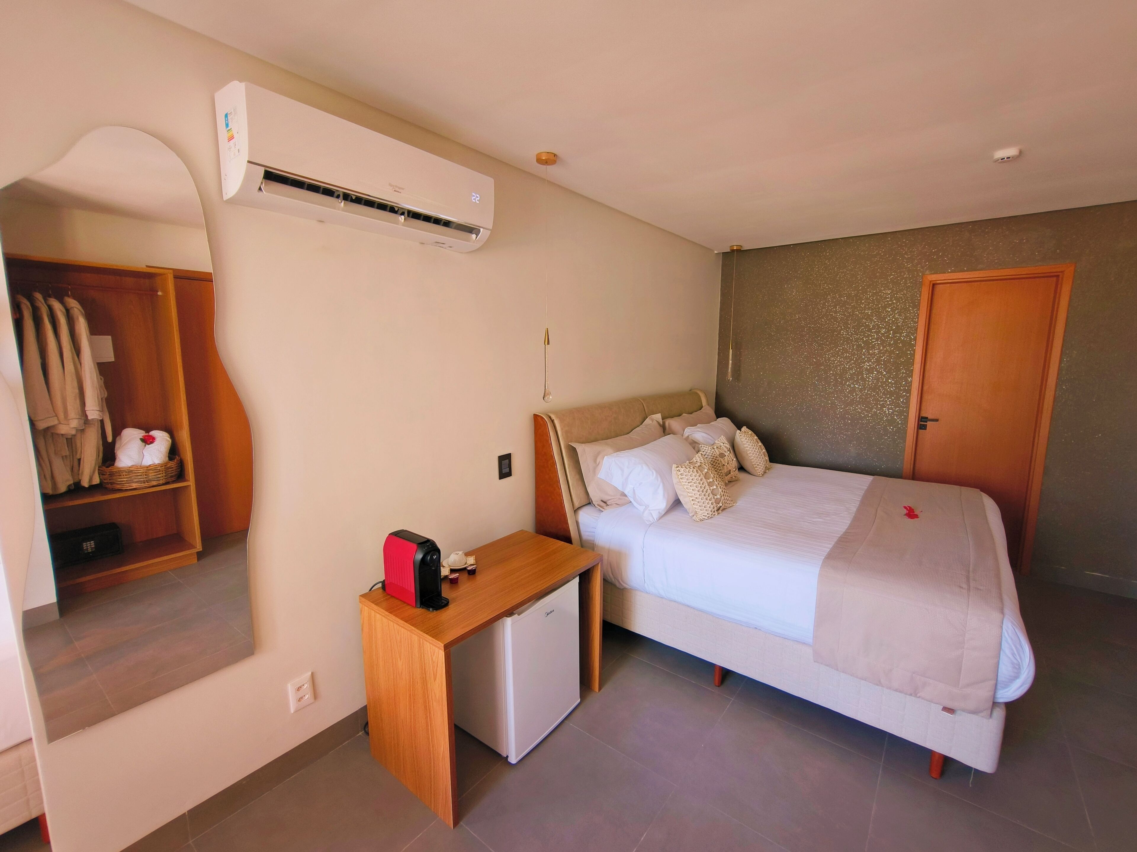 Une chambre avec un lit, une table de chevet et un petit bureau avec un ordinateur. On y trouve également une armoire, un miroir et un appareil de climatisation fixé au mur.