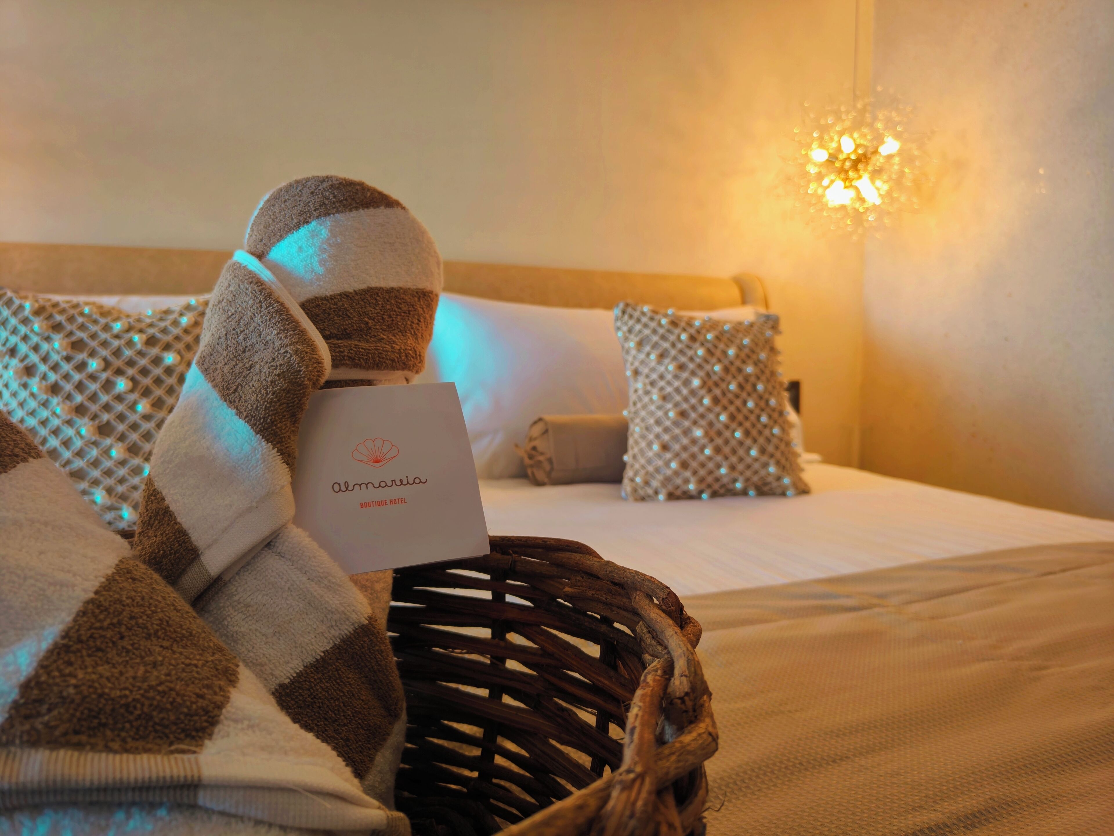 Ein Hotelzimmer mit Bett, Kissen, Decke, einem Korb und einer Karte mit der Aufschrift 'Almaria Boutique Hotel'.
