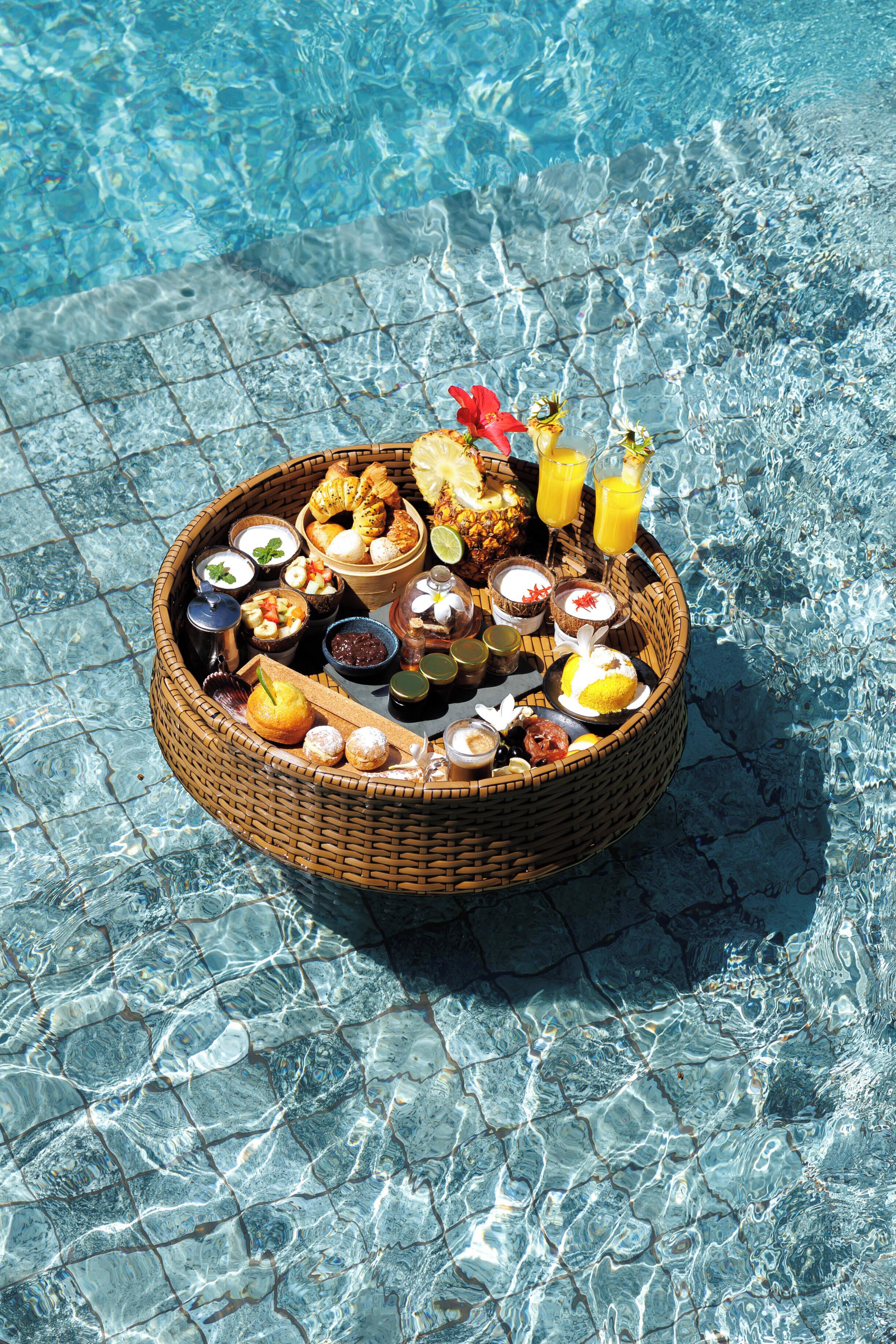 Un panier en osier rempli de divers aliments et boissons, placé sur une surface carrelée près d’une piscine.