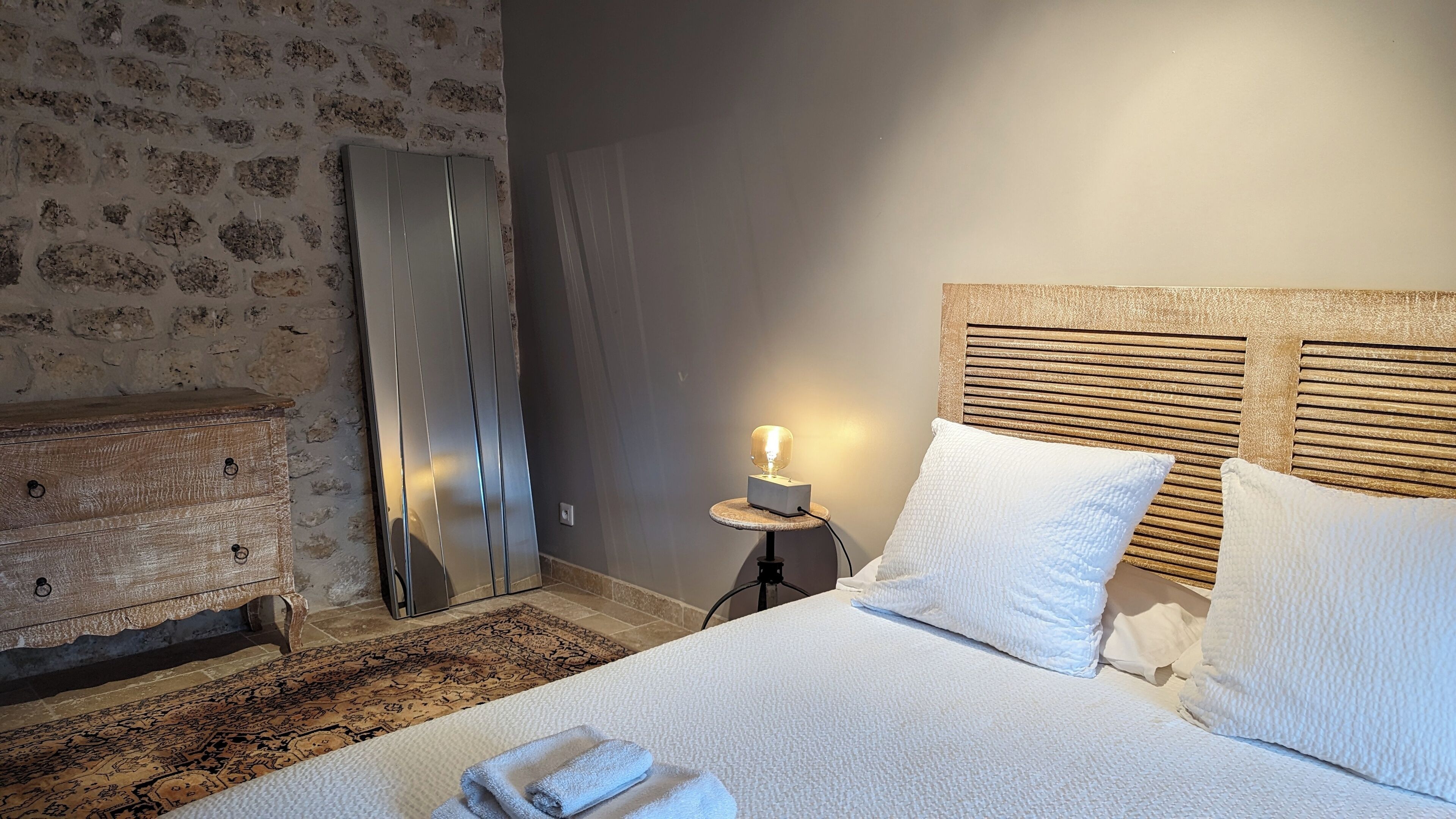 A hotel room with a bed, a wooden dresser, a patterned rug, and a small table with a lamp.