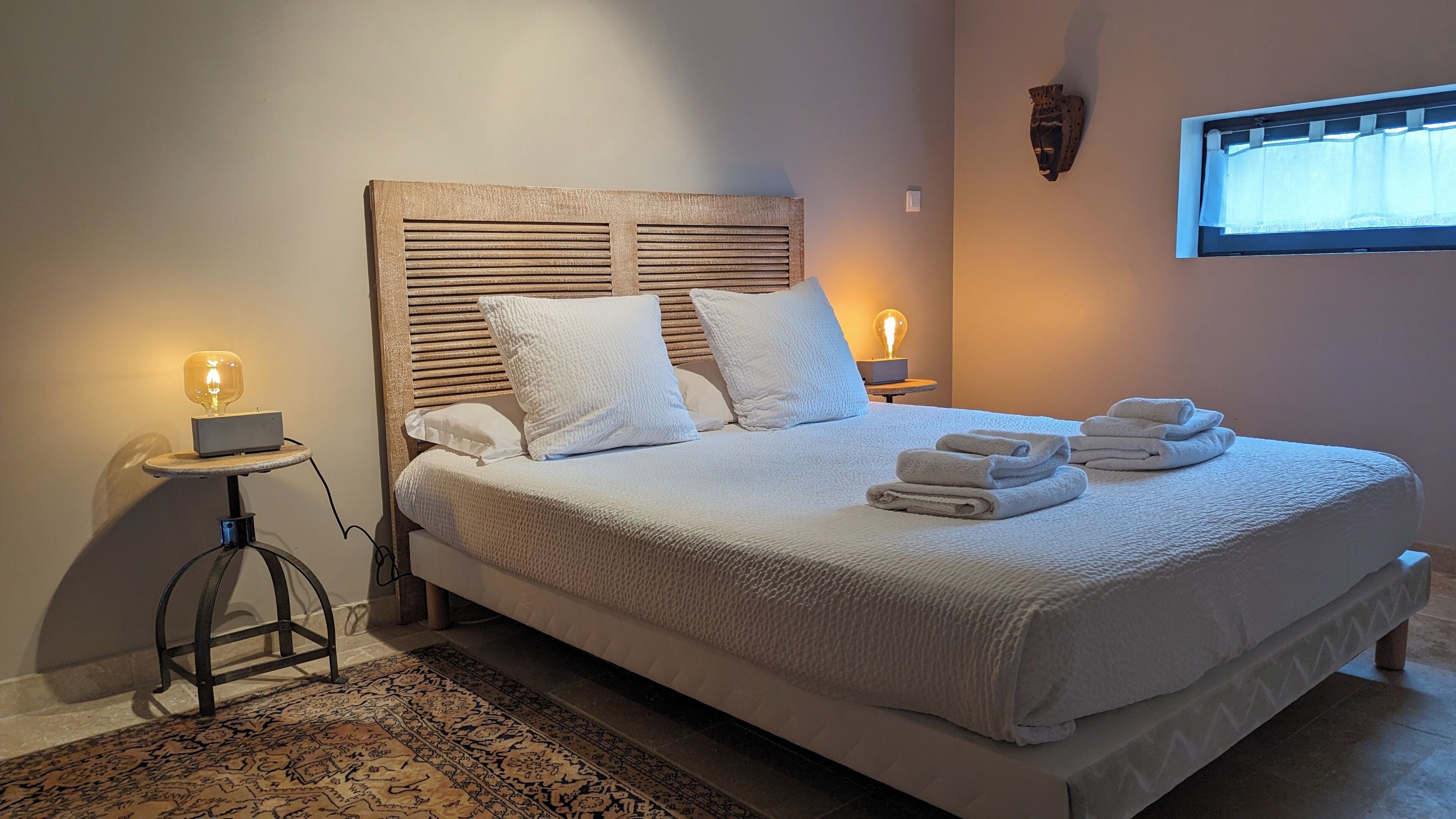 A neatly made bed with a large headboard, two bedside lamps, and a window with a curtain.