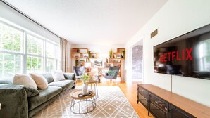 Una sala moderna con un televisor grande mostrando 'NETFLIX', un sofá gris, una mesa de centro y una estantería llena de libros y objetos decorativos.