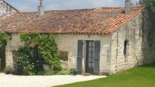 A rustic stone building with a tiled roof, a small garden area with gravel and grass, and a clear sky. - Ground level two-bedroom gîte with gated yard. Ideal for young families. (SOYAUX)