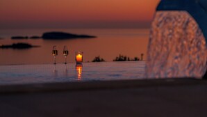Vista al atardecer sobre un cuerpo de agua tranquilo, con dos copas de vino y una vela en una terraza.