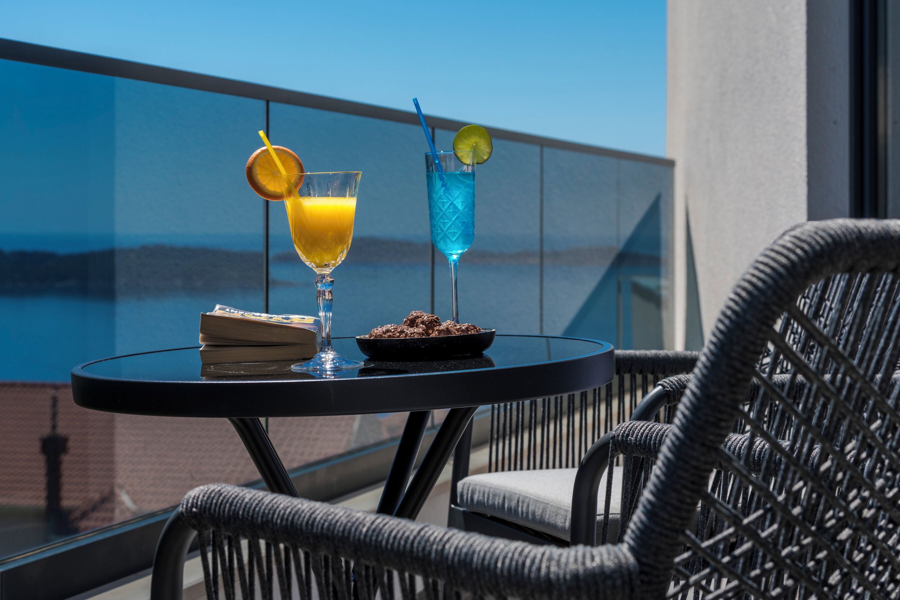 A round table with two drinks, a book, and a plate of food on a balcony overlooking a body of water.