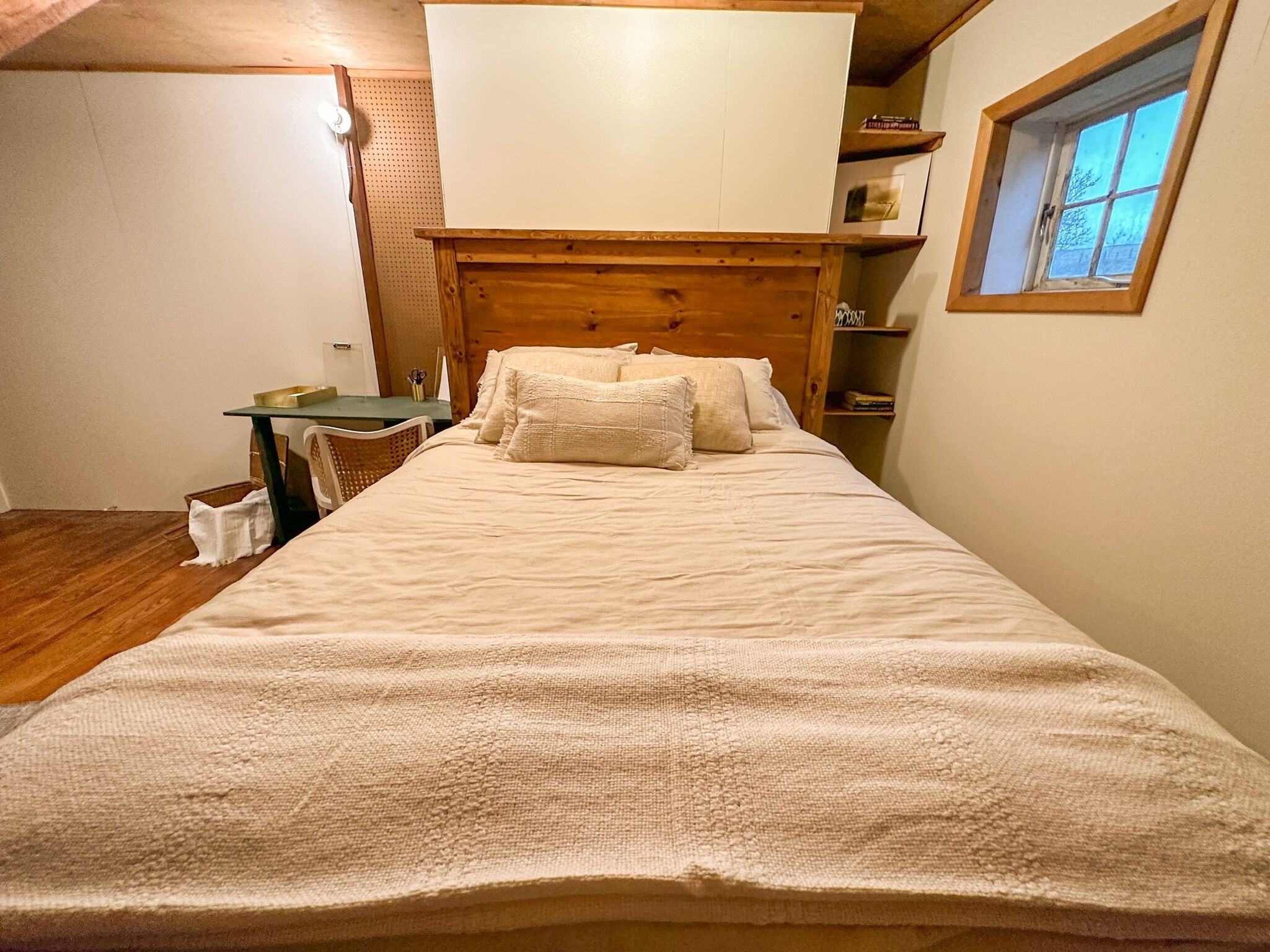 Een slaapkamer met een houten hoofdbord, een bed met wit beddengoed, een tafeltje met een stoel en een raam met uitzicht op bomen.