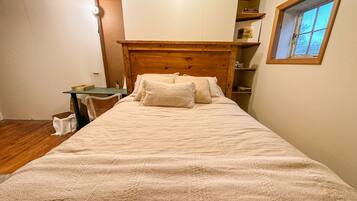 Een slaapkamer met een houten hoofdbord, een bed met wit beddengoed, een tafeltje met een stoel en een raam met uitzicht op bomen.