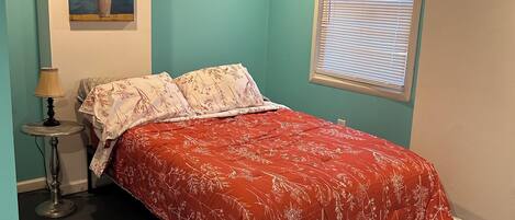 A bedroom with a bed, a nightstand, a lamp, a painting, and a window with blinds.