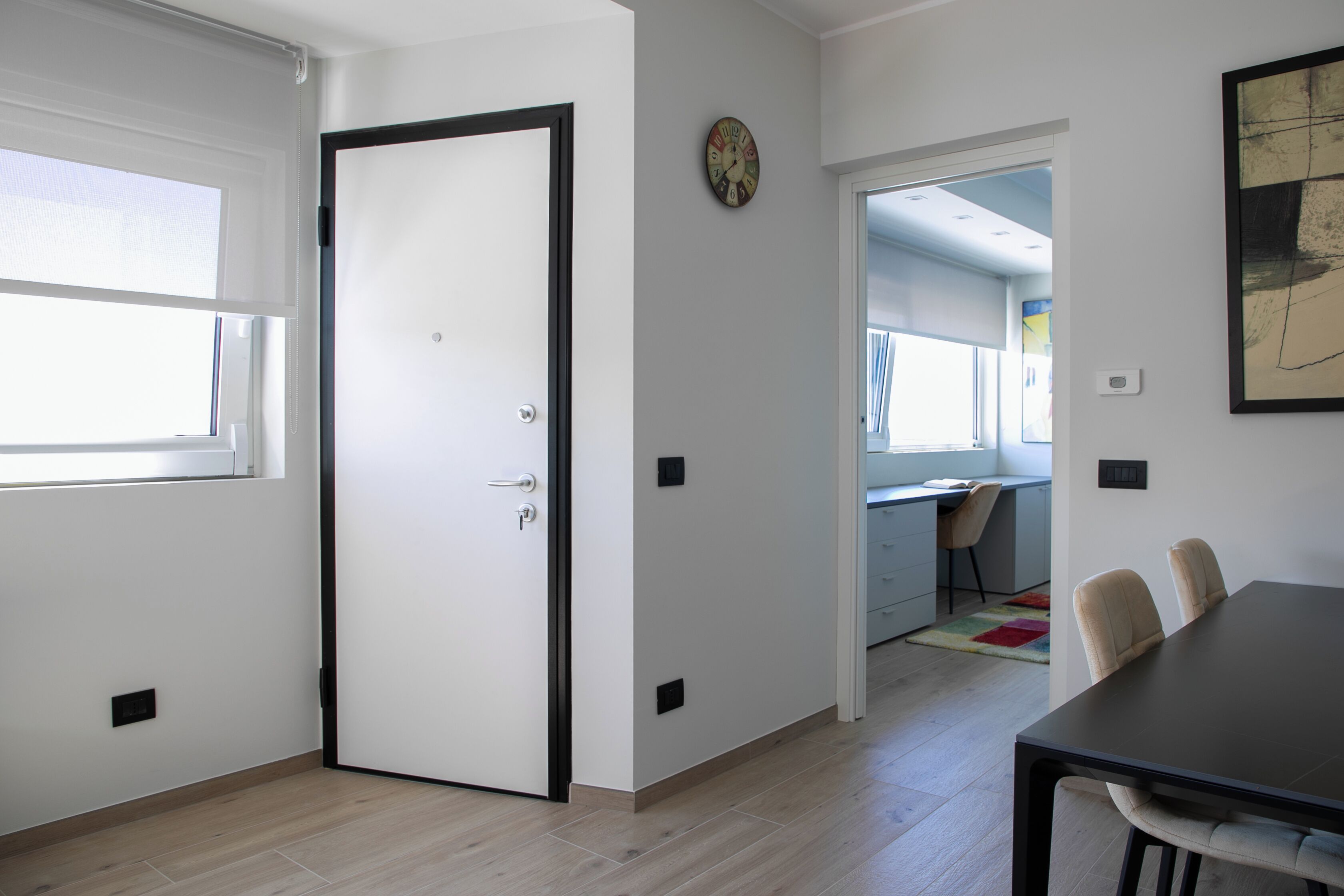 Ein modernes Zimmer mit einem großen Fenster, einer Tür, einem Esstisch mit Stühlen und einer durchgehenden Tür erkennbaren Küchenzeile.