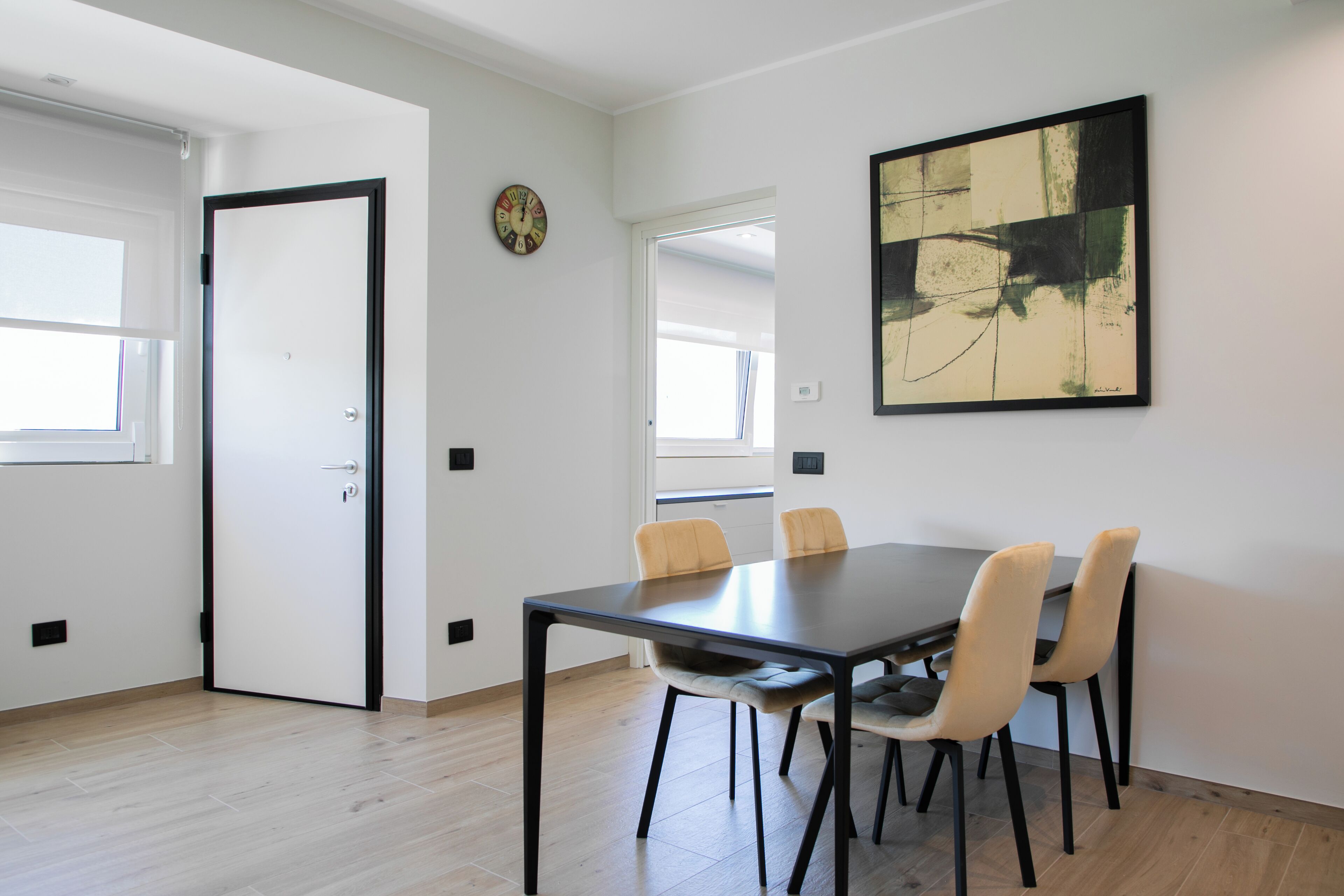A modern dining area with a dark table and four chairs, a clock on the wall, and a large abstract painting.