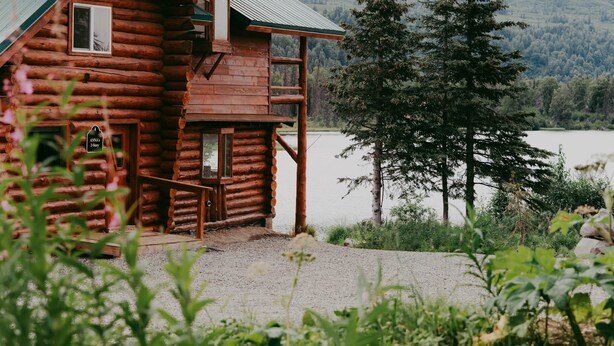 Une cabane en rondins, avec une façade en bois, un toit en métal et une vue sur un lac et une forêt en arrière-plan.