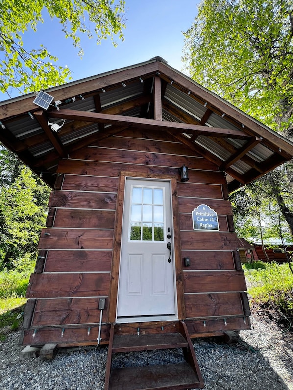 Una cabaña rústica con puerta blanca, un pequeño porche y un cartel que indica que es la 'Cabaña Primitiva #4 Rapha'.