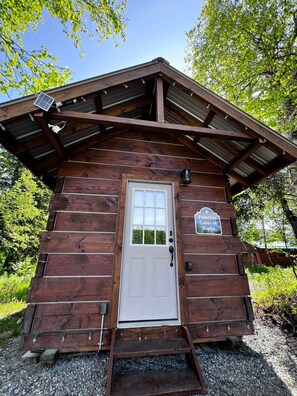 Une cabane rustique avec une porte blanche, un petit porche et une pancarte indiquant quâil sâagit de la « Cabane primitive n° 4, Rapha ».
