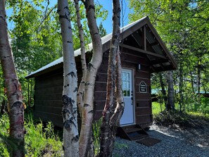 A small cabin with a door and a sign reading 'Primitive Cabin #4 Rapha' in a wooded area.