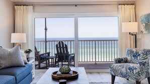 A room with a balcony, a sofa, a chair, a table with a tray, and a lamp. - Seagate 401—Gulf-Front Corner Condo w/ Wraparound Balcony, Pool Access & Sunset Views (Indian Shores)