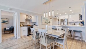 A modern kitchen with white cabinetry, a central island, pendant lighting, and a dining area with white chairs. - Seagate 401—Gulf-Front Corner Condo w/ Wraparound Balcony, Pool Access & Sunset Views (Indian Shores)