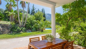 A wooden table and chairs under a white pergola, surrounded by lush greenery and a clear view of a swimming pool.