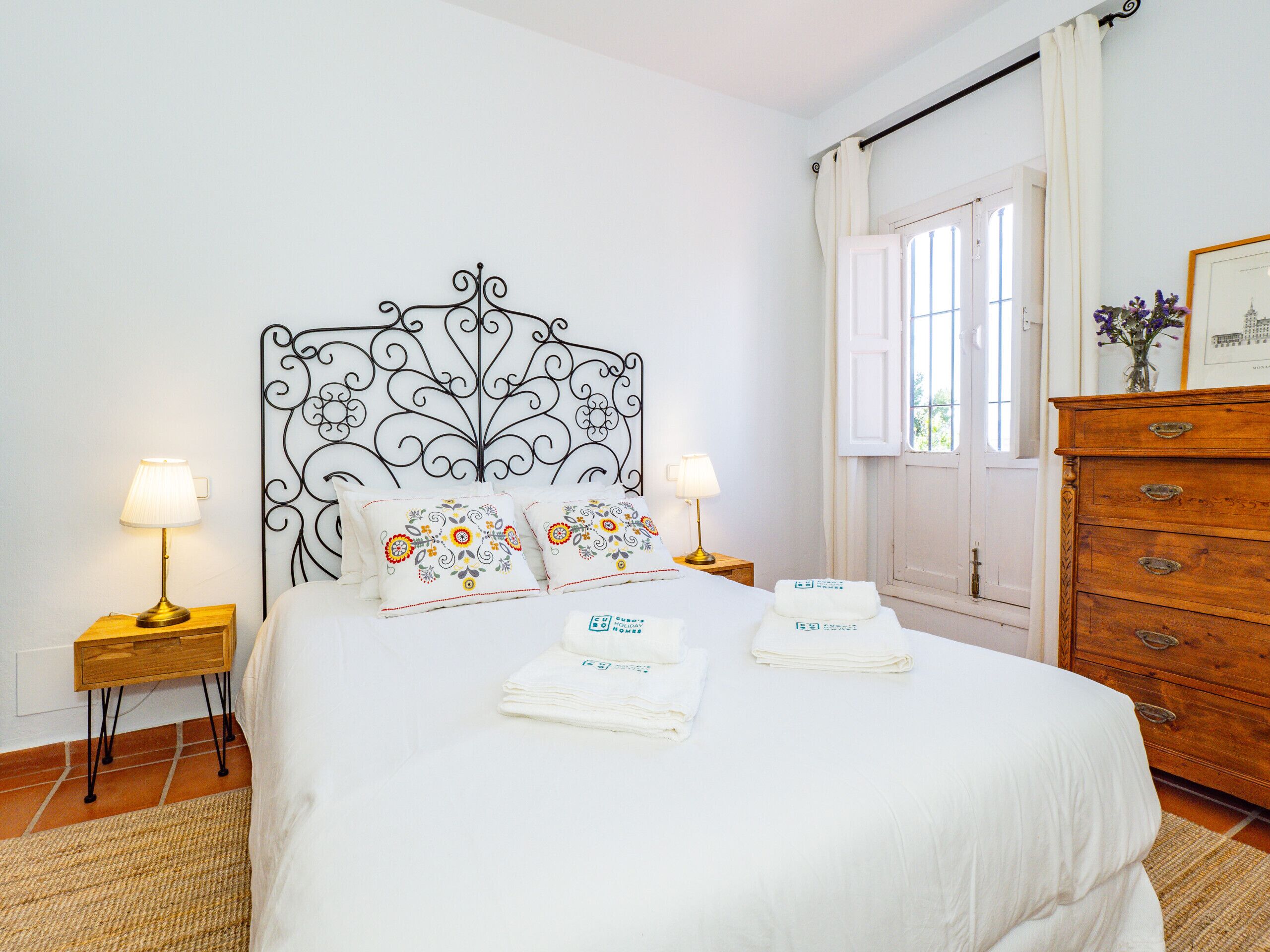 A neatly made bed with decorative pillows, a bedside lamp, and a wooden dresser in a room with white walls and a window.