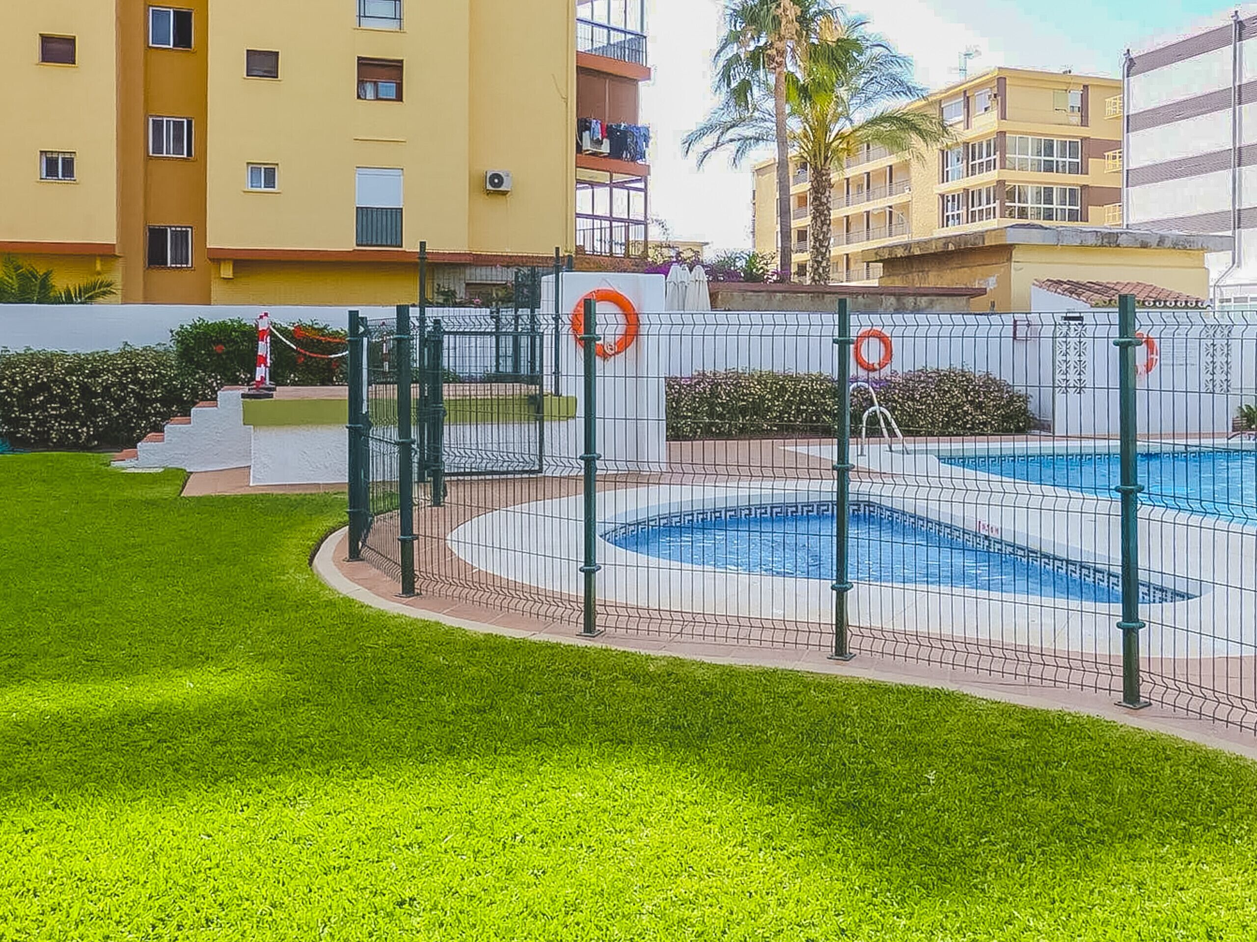 Une piscine entourée d’une clôture, avec des bouées de sauvetage à proximité, une pelouse et un immeuble à plusieurs étages en arrière-plan.