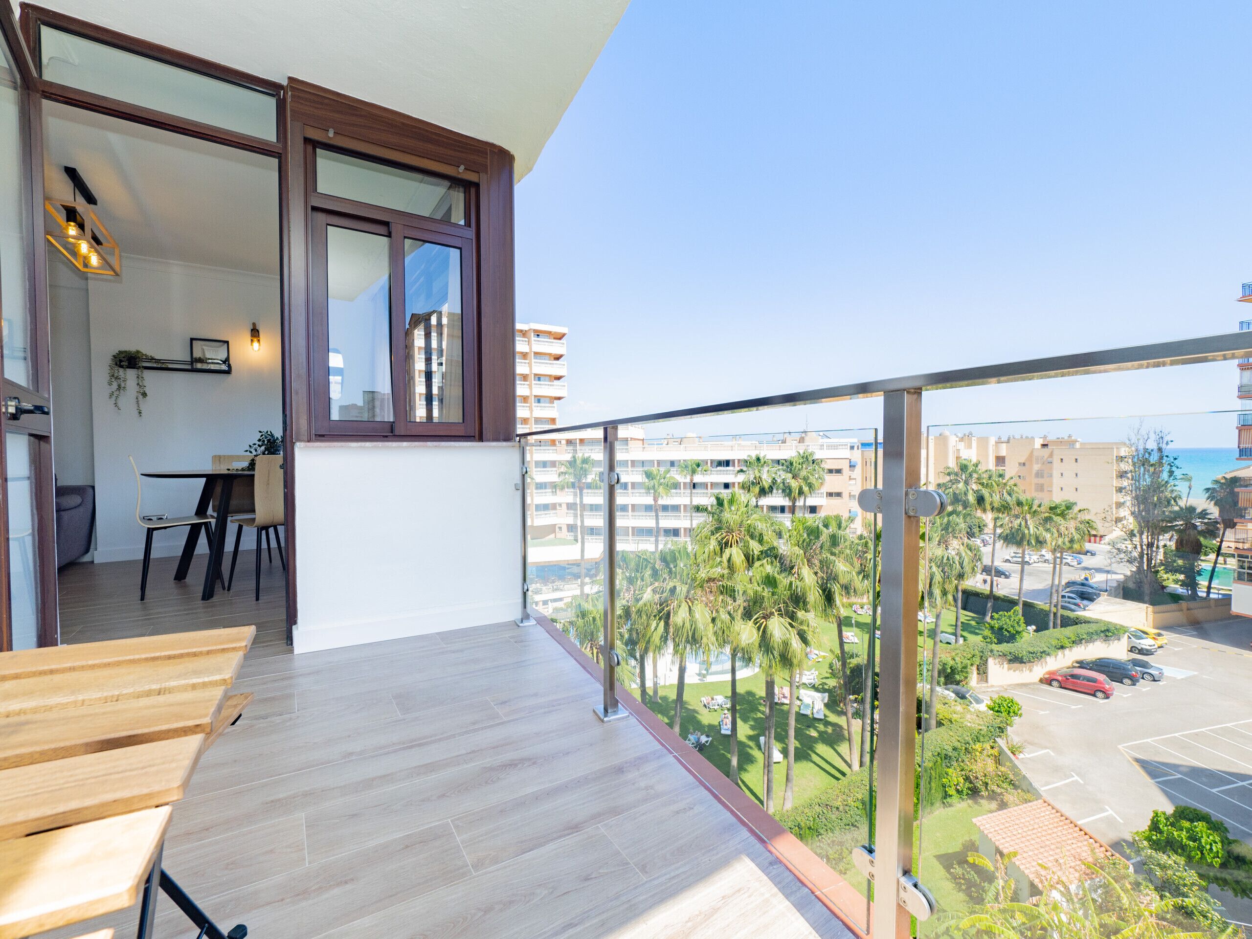 Een balkon met houten vloer, een zithoek met tafel en stoelen, en uitzicht op een parkeerplaats en gebouwen.