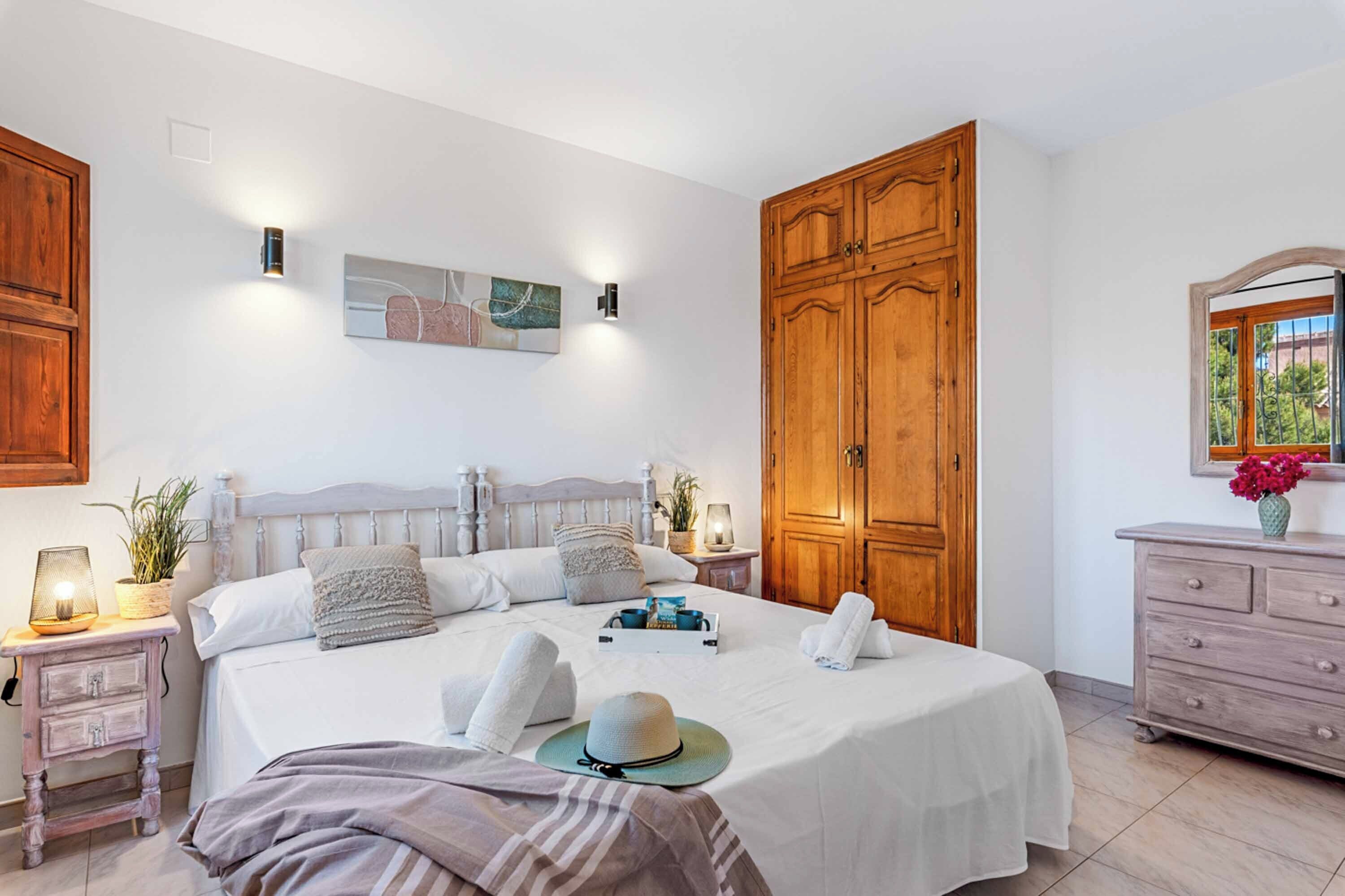 A bedroom with a large bed, wooden wardrobe, bedside tables with lamps, and a dresser with a vase of flowers.