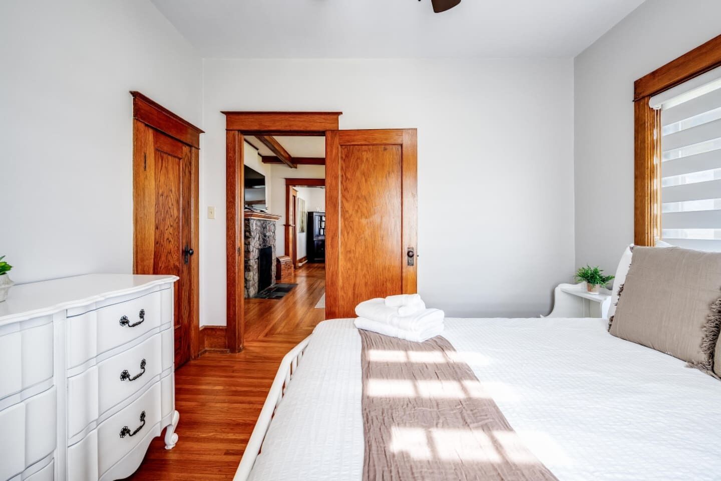 Una camera da letto con pavimento in legno, mobili bianchi, un letto con copriletto bianco e vista su un corridoio con camino.