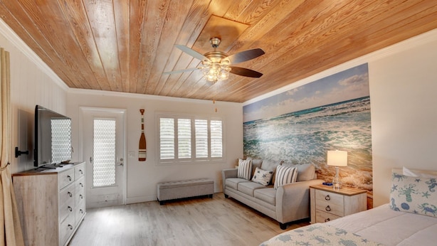 A bedroom with a wooden ceiling, a ceiling fan, a television, a bench, a sofa, a bed, and a mural of the ocean. - Beach Bungalow  (Delray Beach)
