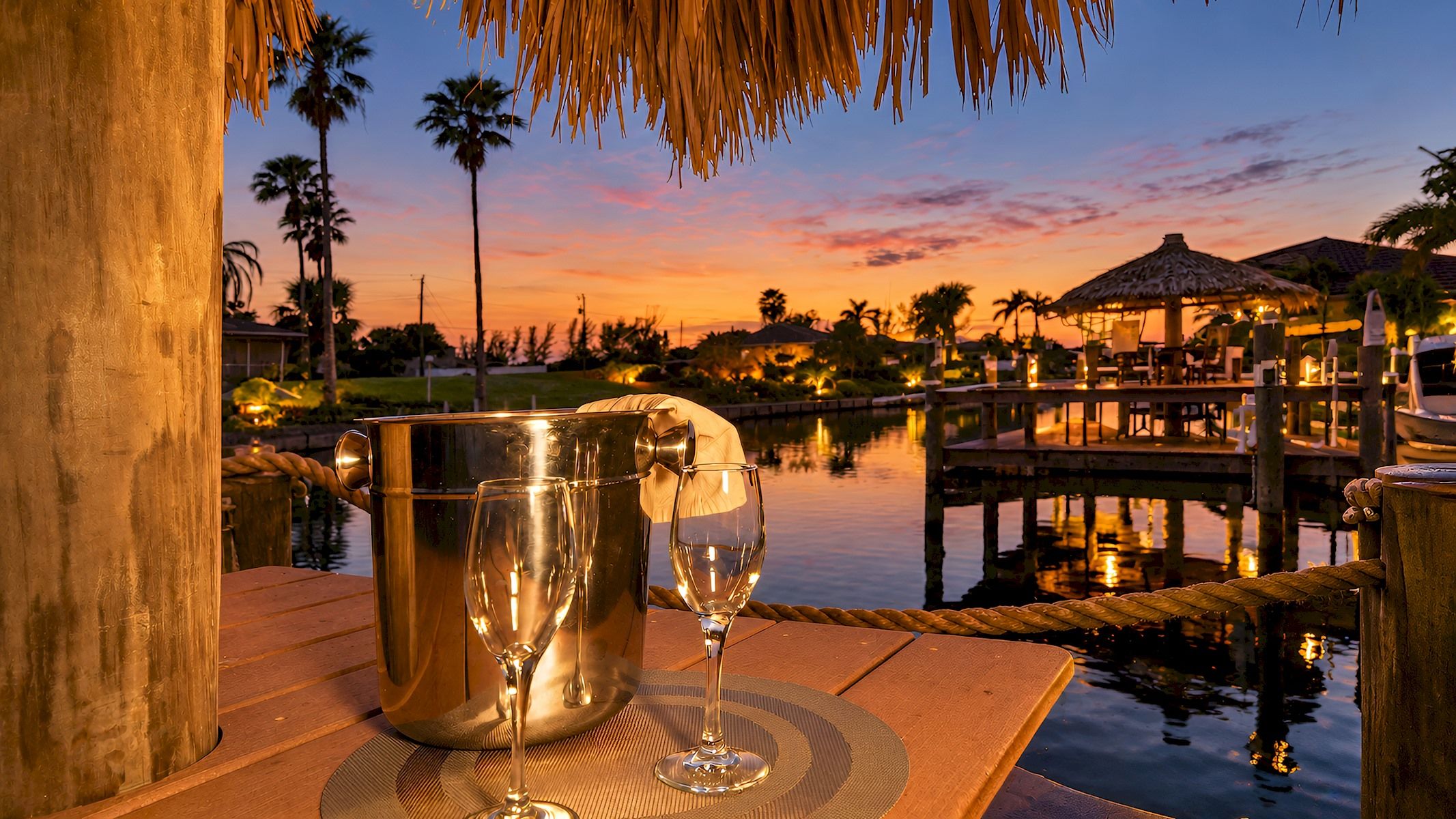 A sunset view with a wooden dock, a thatched-roof structure, and a calm body of water.